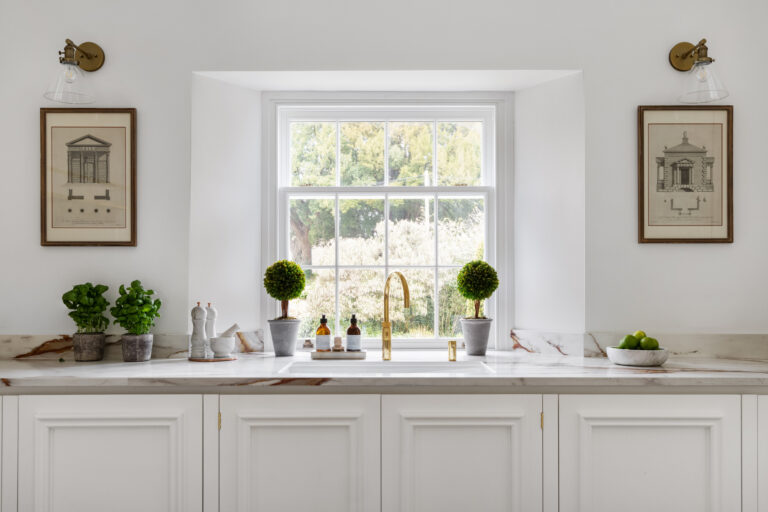 White cabinetry beneath a sash window, with a brass tap, veined worktop and topiary either side of the sink