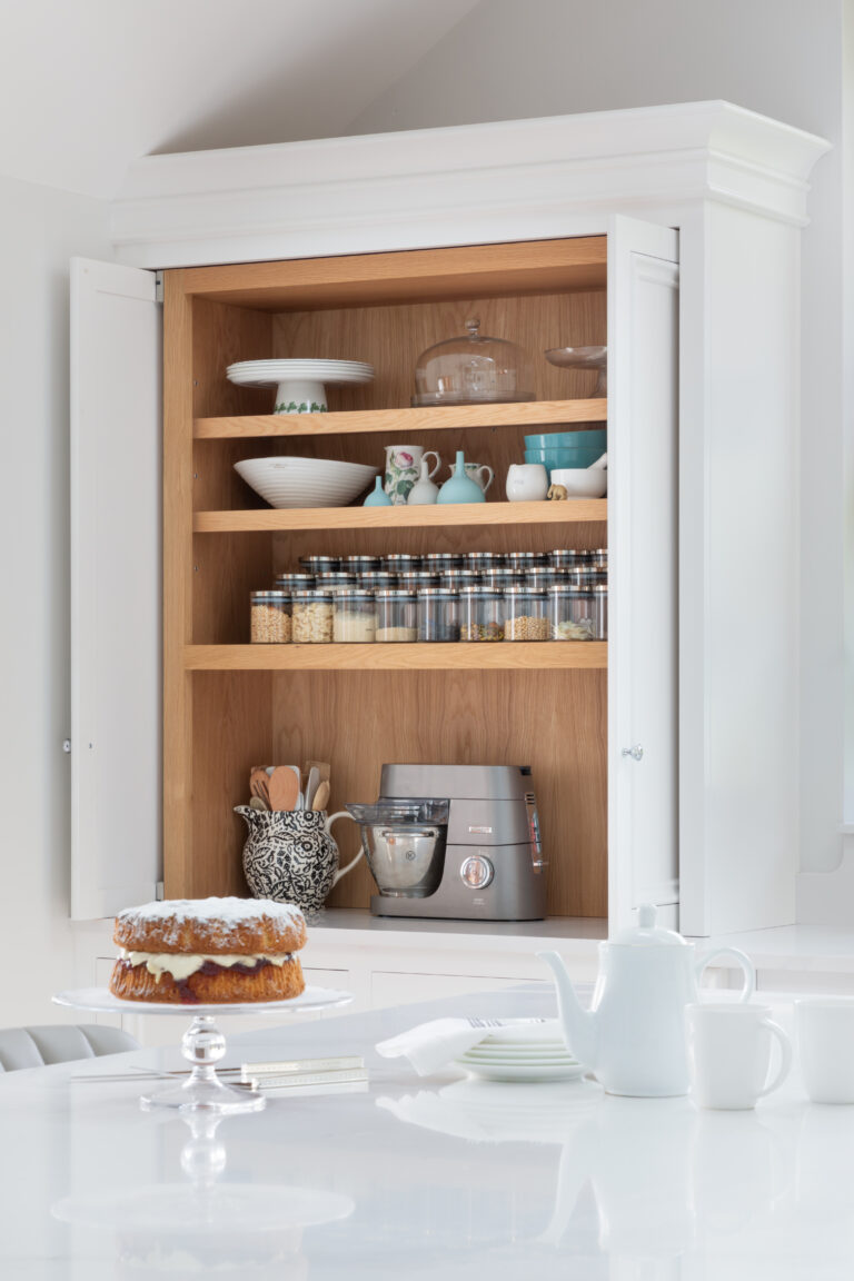 Open pantry cupboard with timber-lined shelving storing crockery, storage jars and a stand mixer, with a cake on the worktop below