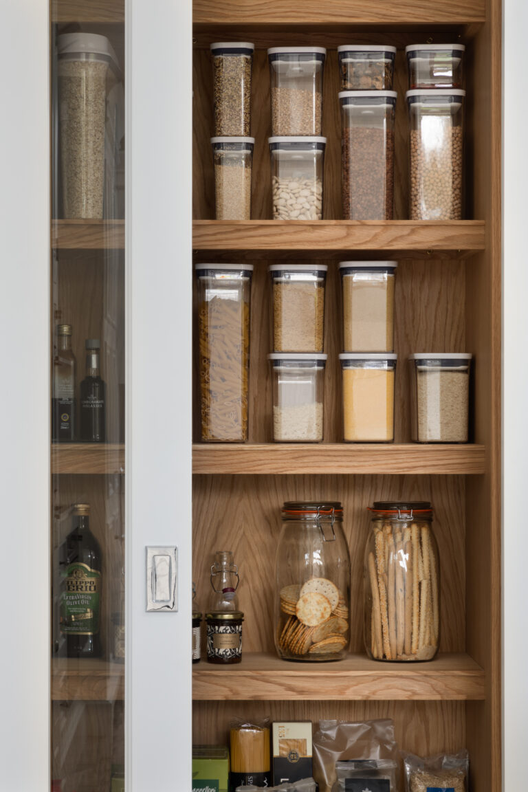 Walk-in pantry shelves lined with glass containers of dry goods, jars and bottles, behind a glazed cupboard door