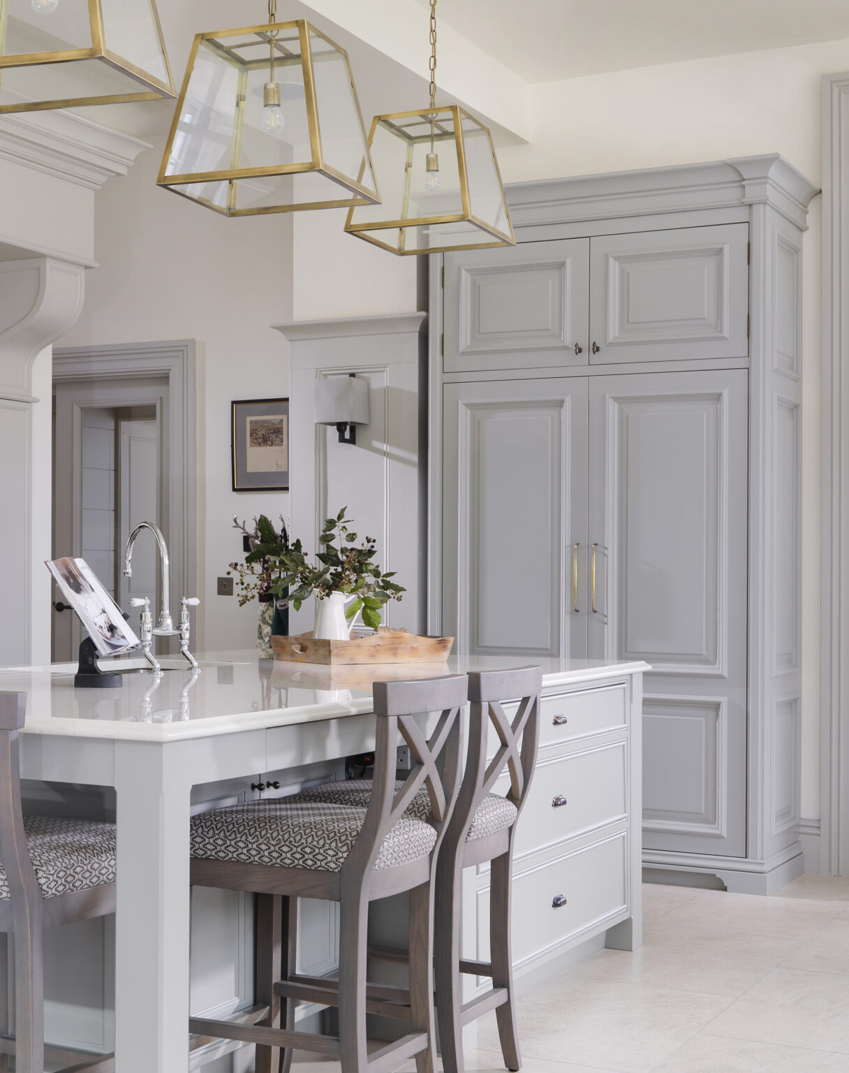 Grey panelled kitchen island with brass pendant lanterns, tall storage cabinetry and upholstered bar stools