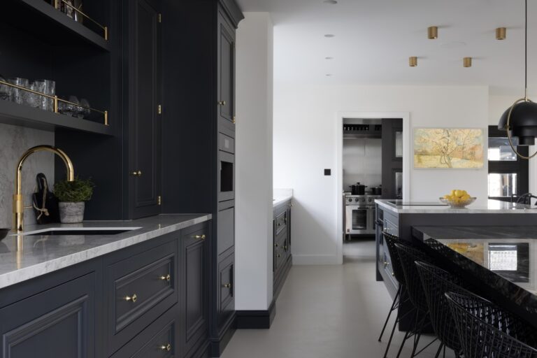 Dark cabinetry with brass handles and tap, stone worktop, and a view through to the hidden prep kitchen beyond
