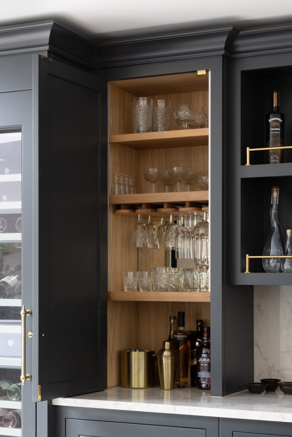 Open bar cabinet with timber-lined shelving storing crystal glassware, spirit bottles and brass cocktail accessories