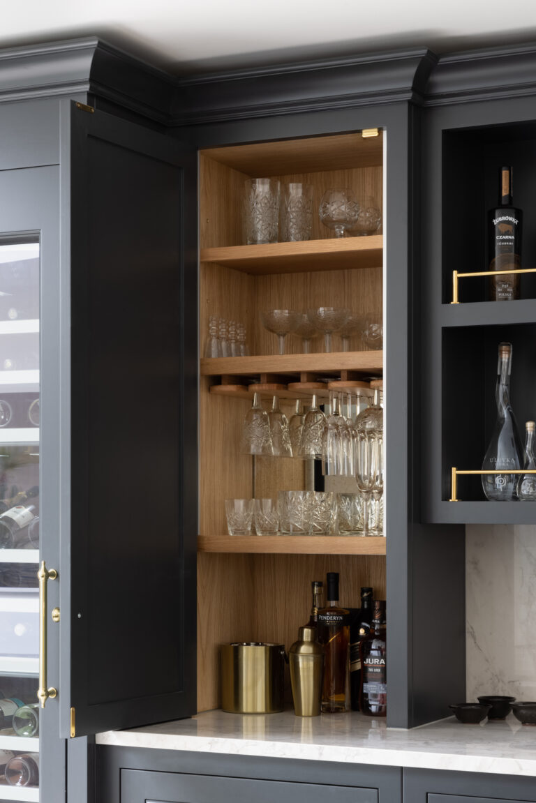 Open bar cabinet with timber-lined shelving storing crystal glassware, spirit bottles and brass cocktail accessories