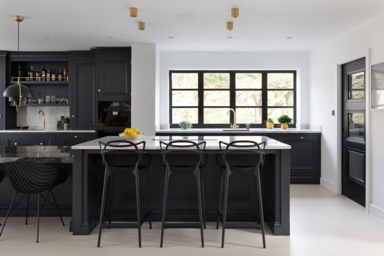 Kitchen with dark cabinetry, XTONE Orobico Dark stone island, brass fittings and black bar stools against large windows