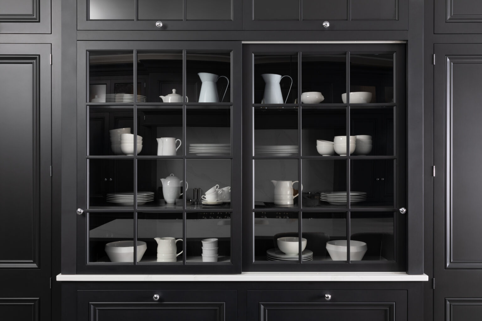 Tall glazed dresser with sliding glass doors displays white crockery against dark cabinetry, with white stone worktop below