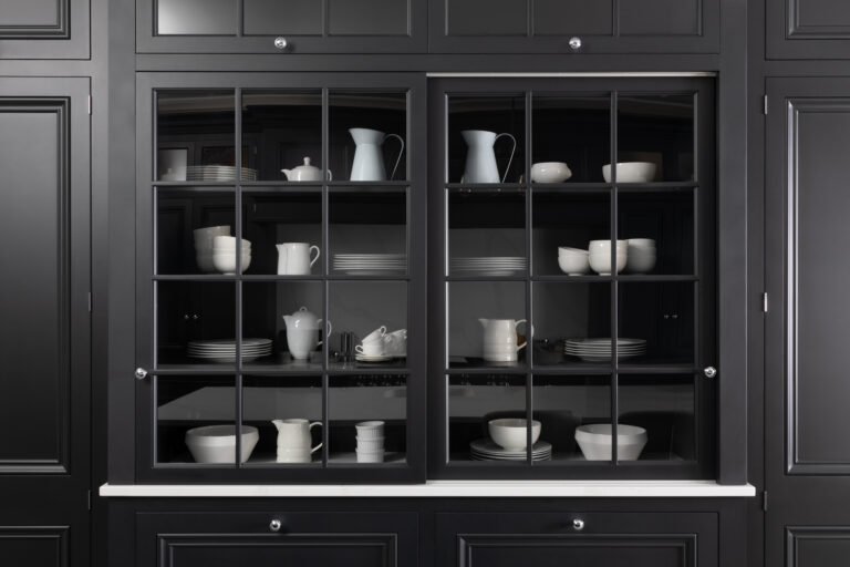 Tall glazed dresser with sliding glass doors displays white crockery against dark cabinetry, with white stone worktop below