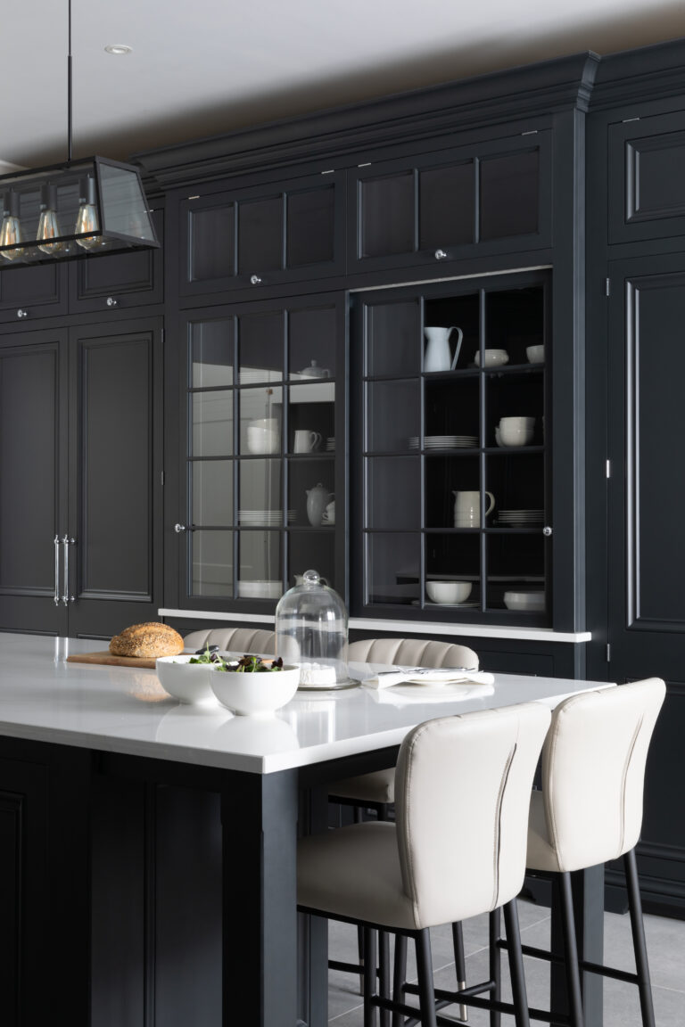 Black kitchen with a tall glazed dresser displaying crockery, white stone island worktop and upholstered bar stools