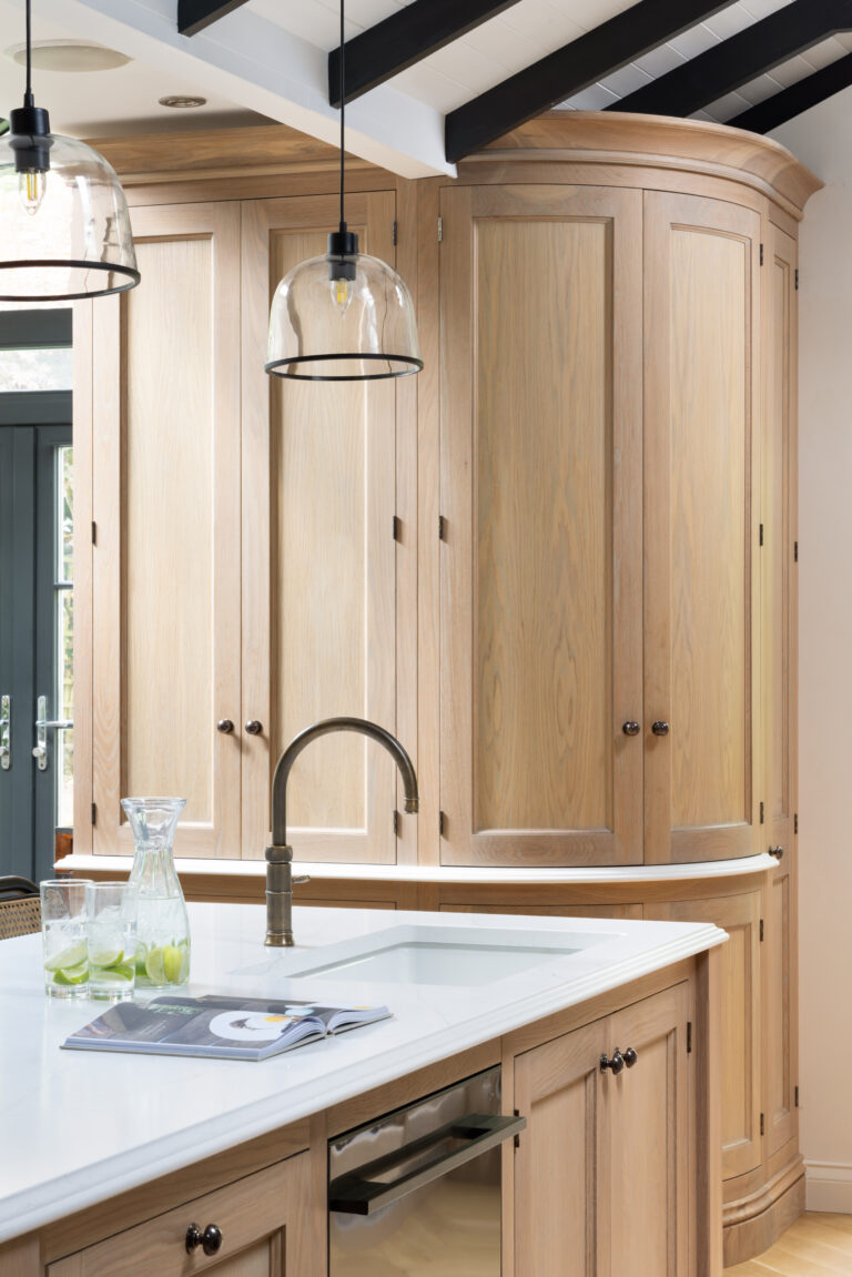 Curved timber cabinetry with antique brass tap and handles, glass pendant lights hanging above a white worktop sink