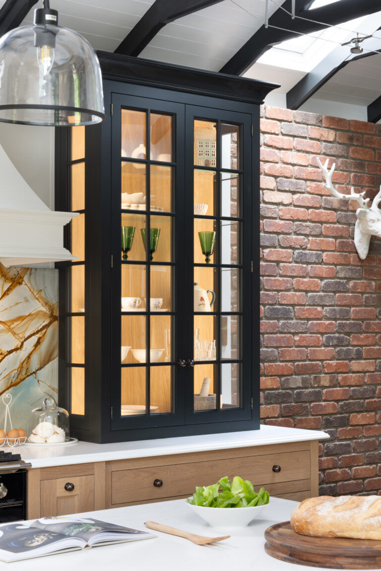 Glazed dark display cabinet with glass-panelled doors showing crockery, set above timber drawers against an exposed brick wall