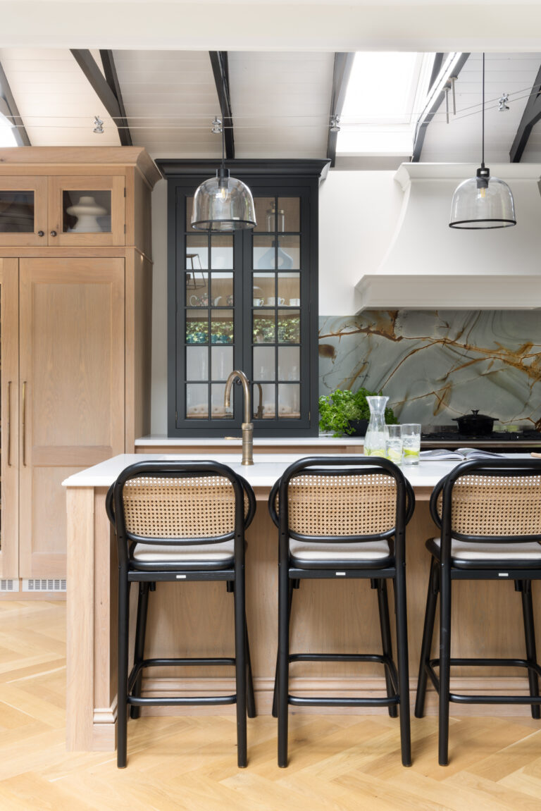 Kitchen island with cane-backed bar stools, antique brass tap, and backlit stone splashback behind glazed dark cabinetry