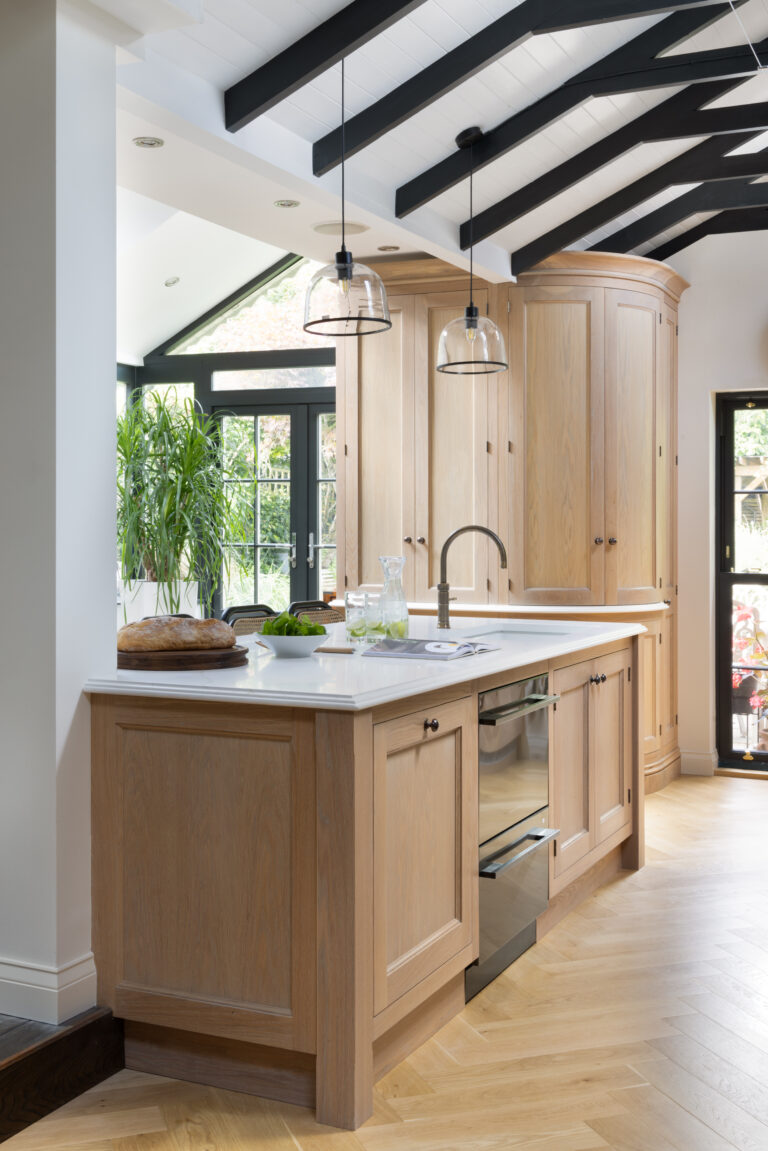 Kitchen island with pale timber cabinetry, white worktop and curved corner units beneath vaulted beamed ceiling