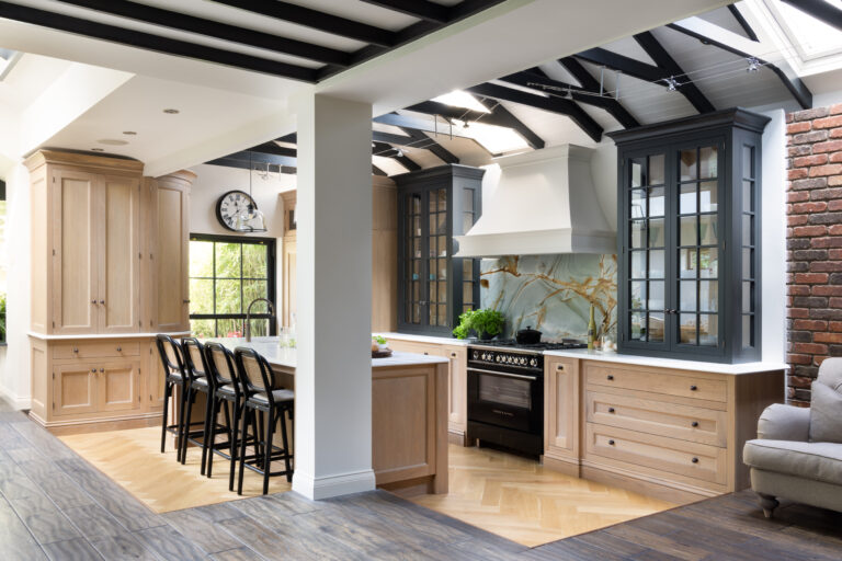 Open-plan kitchen with vaulted beamed ceiling, pale timber cabinetry, dark display cupboards and a backlit stone splashback behind the range cooker