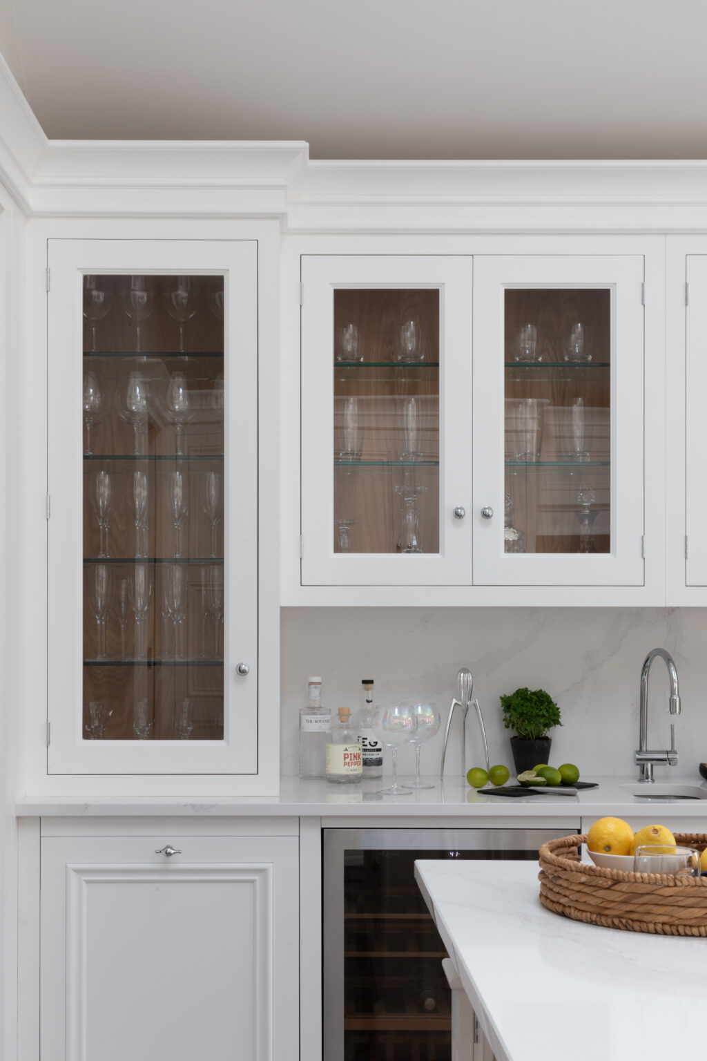 Glass-fronted white cabinets display glassware above a worktop with spirits, a built-in wine cooler and chrome tap