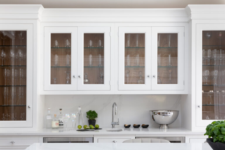 White glass-fronted wall cabinets display glassware above a wet bar with a mixer tap and bottles
