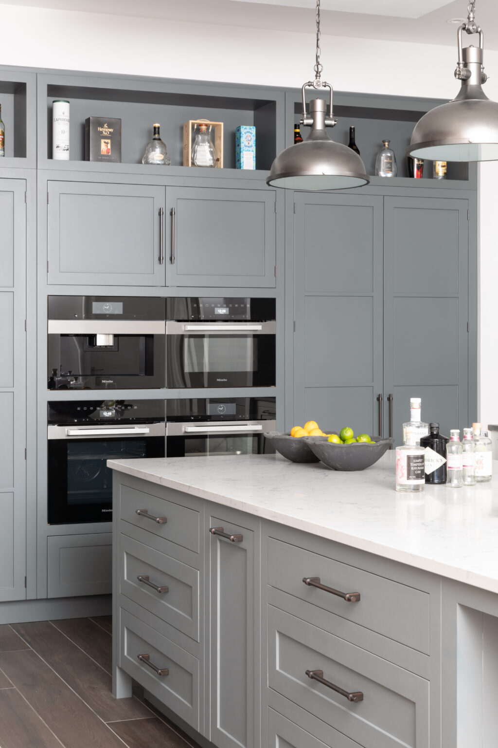 Grey bespoke cabinetry with bronze handles, integrated Miele appliances, open shelving and an island with industrial pendant lights