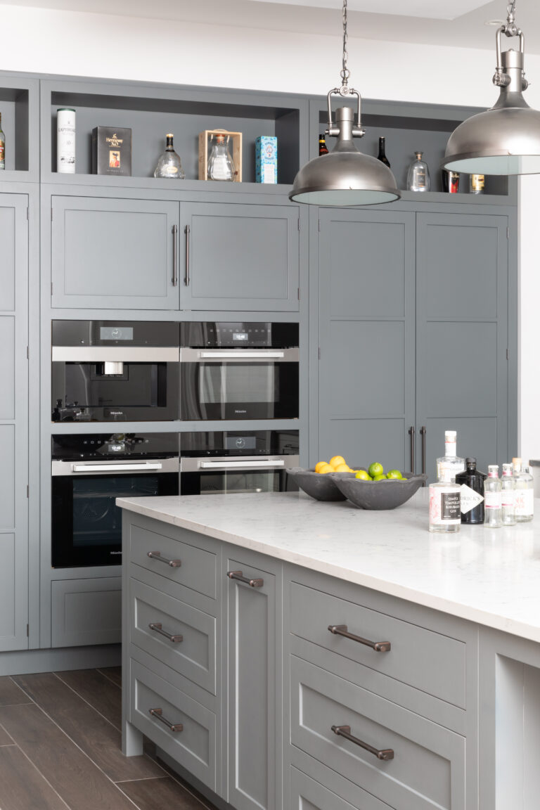 Grey bespoke cabinetry with bronze handles, integrated Miele appliances, open shelving and an island with industrial pendant lights