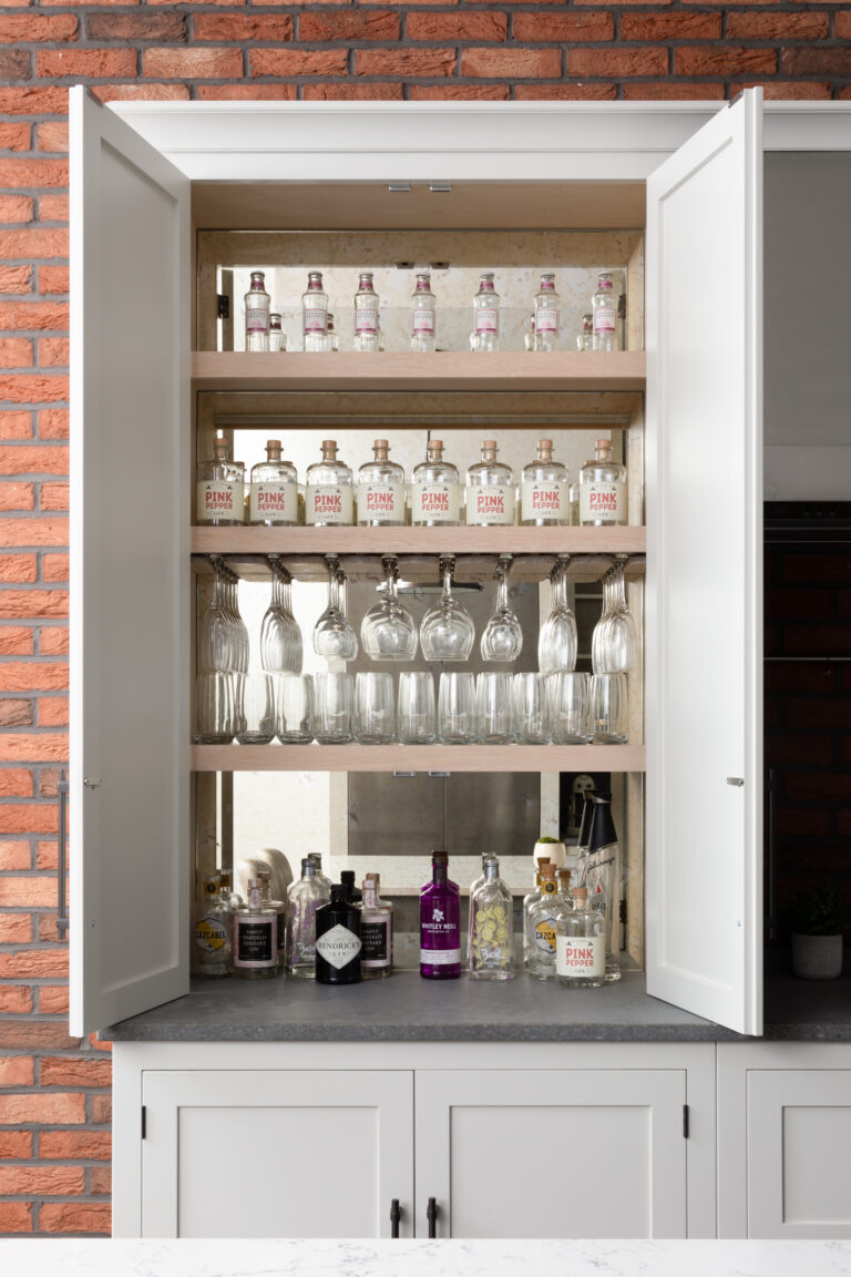 Open drinks cabinet against exposed brick wall, shelves lined with glassware and gin bottles above a mirrored back panel