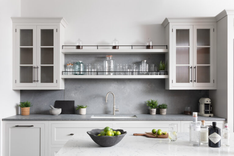 Kitchen sink area with glazed wall cabinets, open shelving holding glassware, and a concrete-effect splashback above the worktop