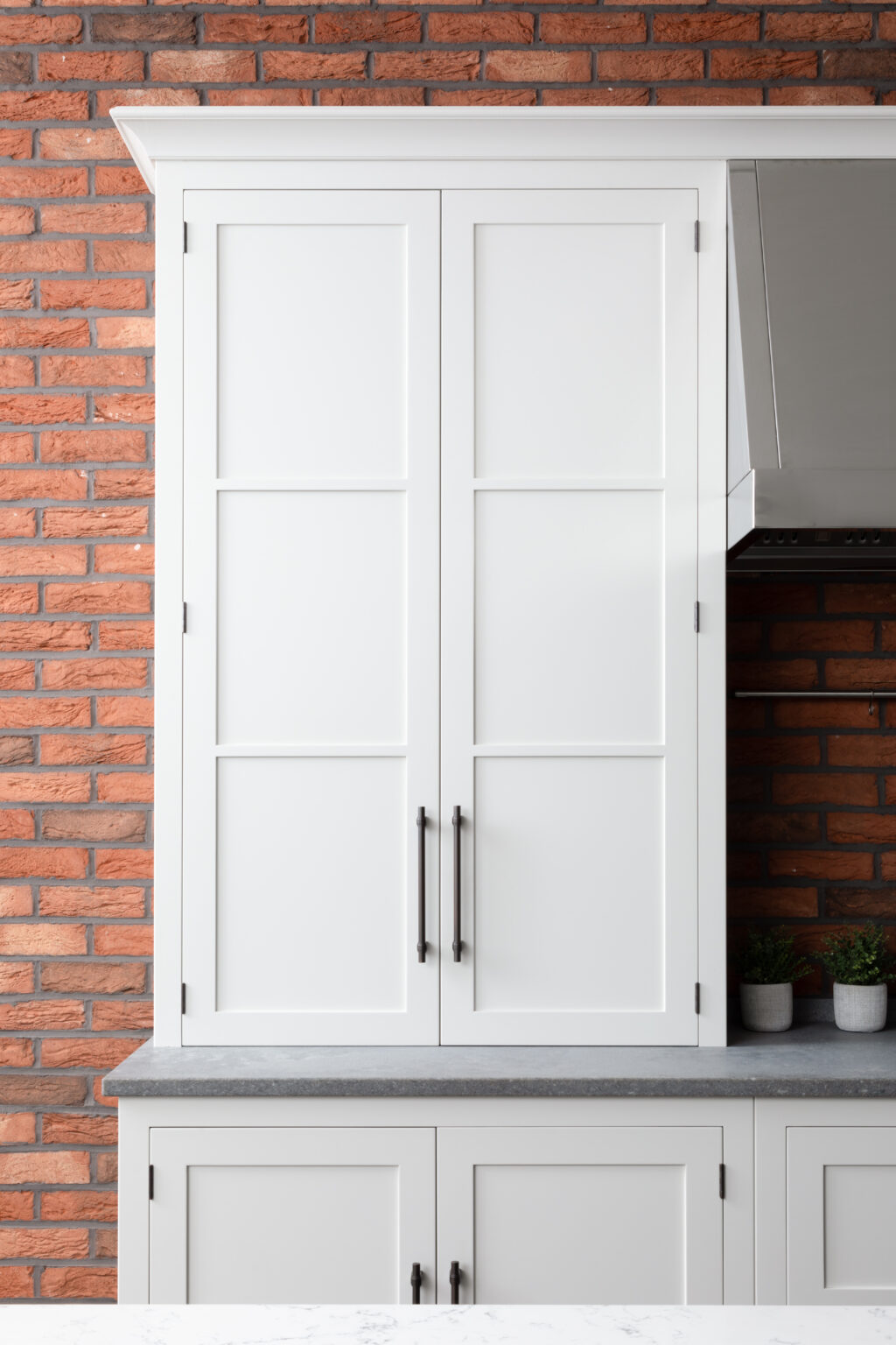 Tall panelled cabinet with bronze handles stands against exposed brick, beside a stainless steel extractor hood