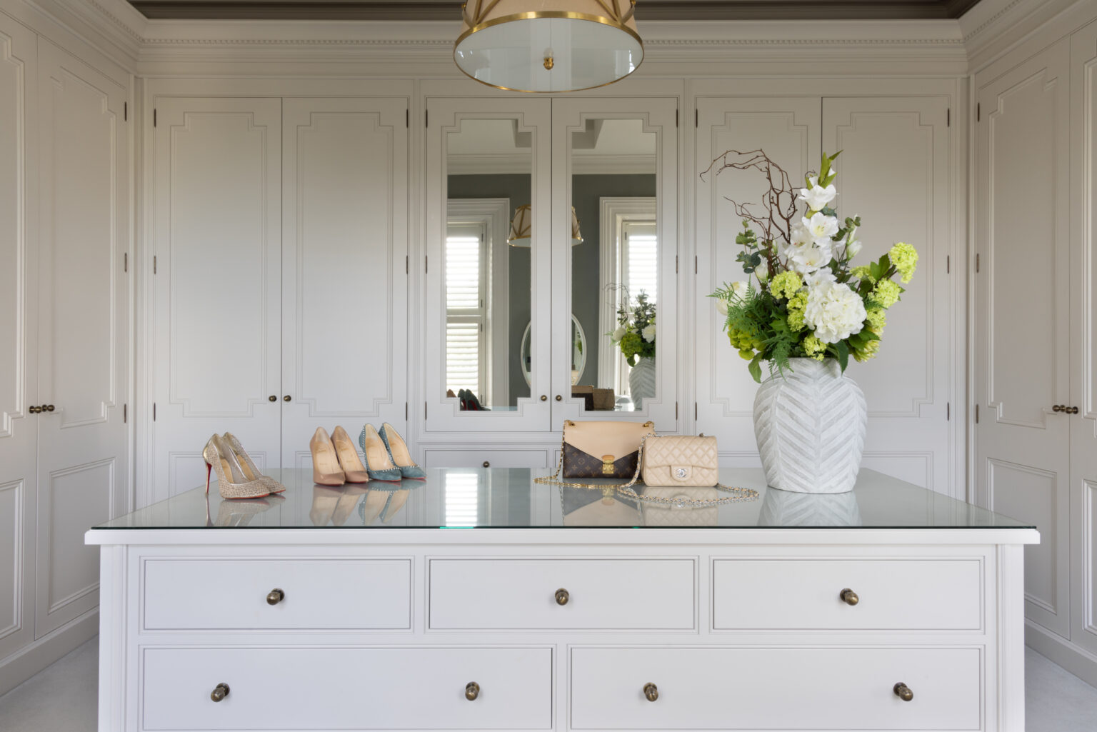 Dressing room island with glass top, fitted wardrobes, displayed shoes, handbags and a flower arrangement