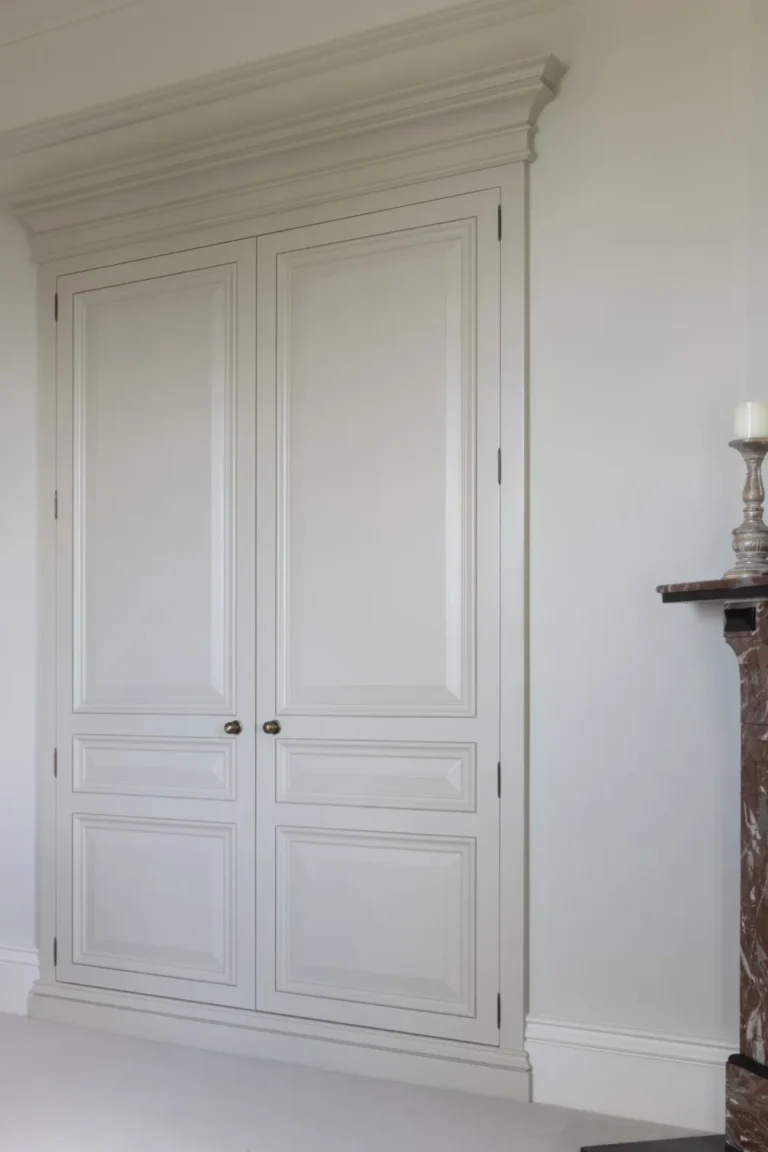 Floor-to-ceiling fitted wardrobe in soft off-white with panelled doors and a substantial cornice beside a stone fireplace