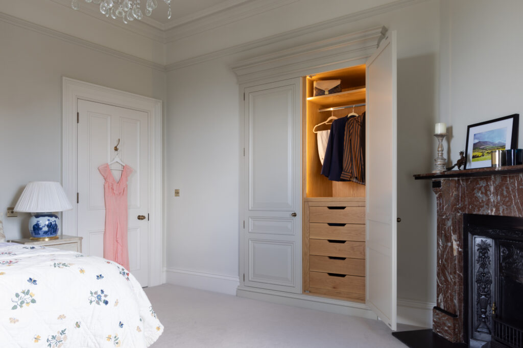 Bedroom with a fitted panelled wardrobe featuring internal timber drawers, hanging rail and a traditional fireplace alongside
