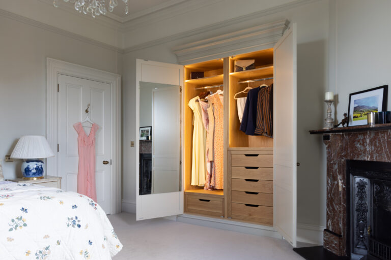 Bespoke bedroom wardrobe with internal lighting, hanging rails, drawers and a mirrored door beside a period fireplace