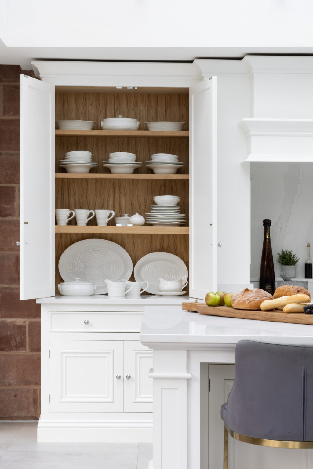 White painted dresser with open doors reveals white crockery on timber-lined shelves beside an exposed sandstone wall