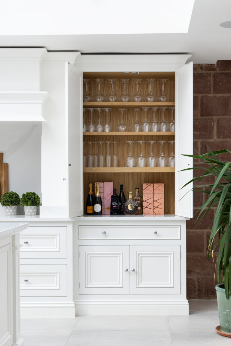 Open white cabinet with internal wood-lined shelves displaying wine glasses and bottled drinks, beside an exposed sandstone wall