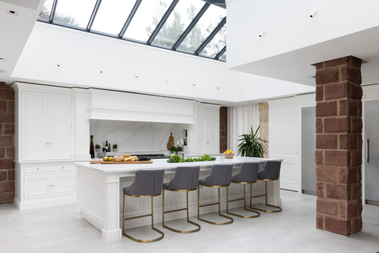 White kitchen island with grey upholstered stools, set beneath a skylight beside an exposed sandstone wall
