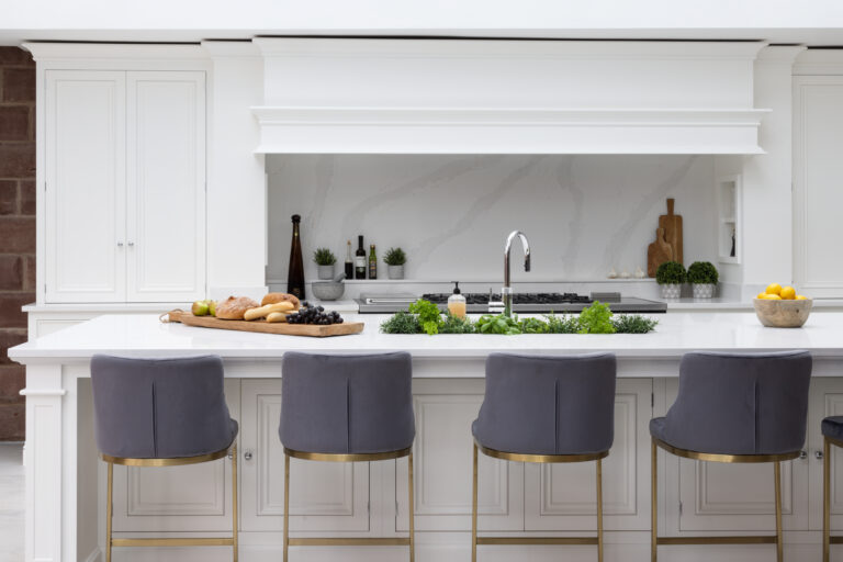 White kitchen island with grey upholstered stools, hob, chrome tap and herbs set against exposed sandstone wall