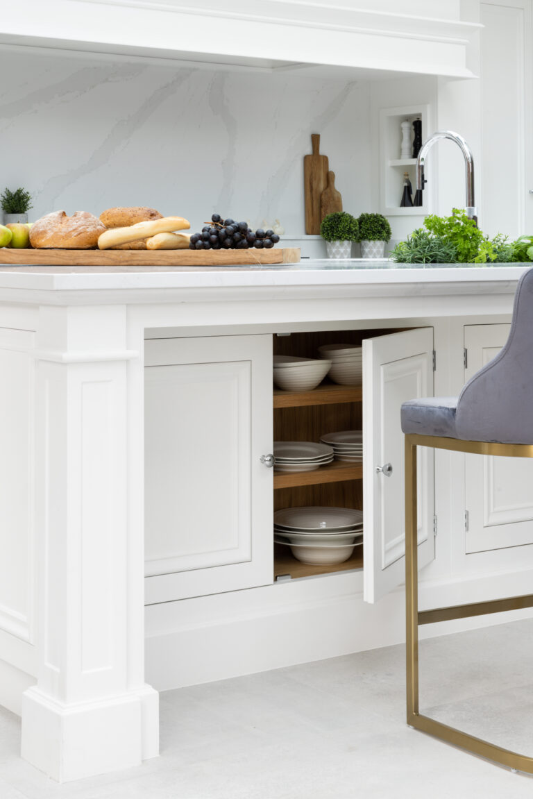 White kitchen island with an open cupboard revealing crockery, plus a worktop styled with bread, grapes and fresh herbs