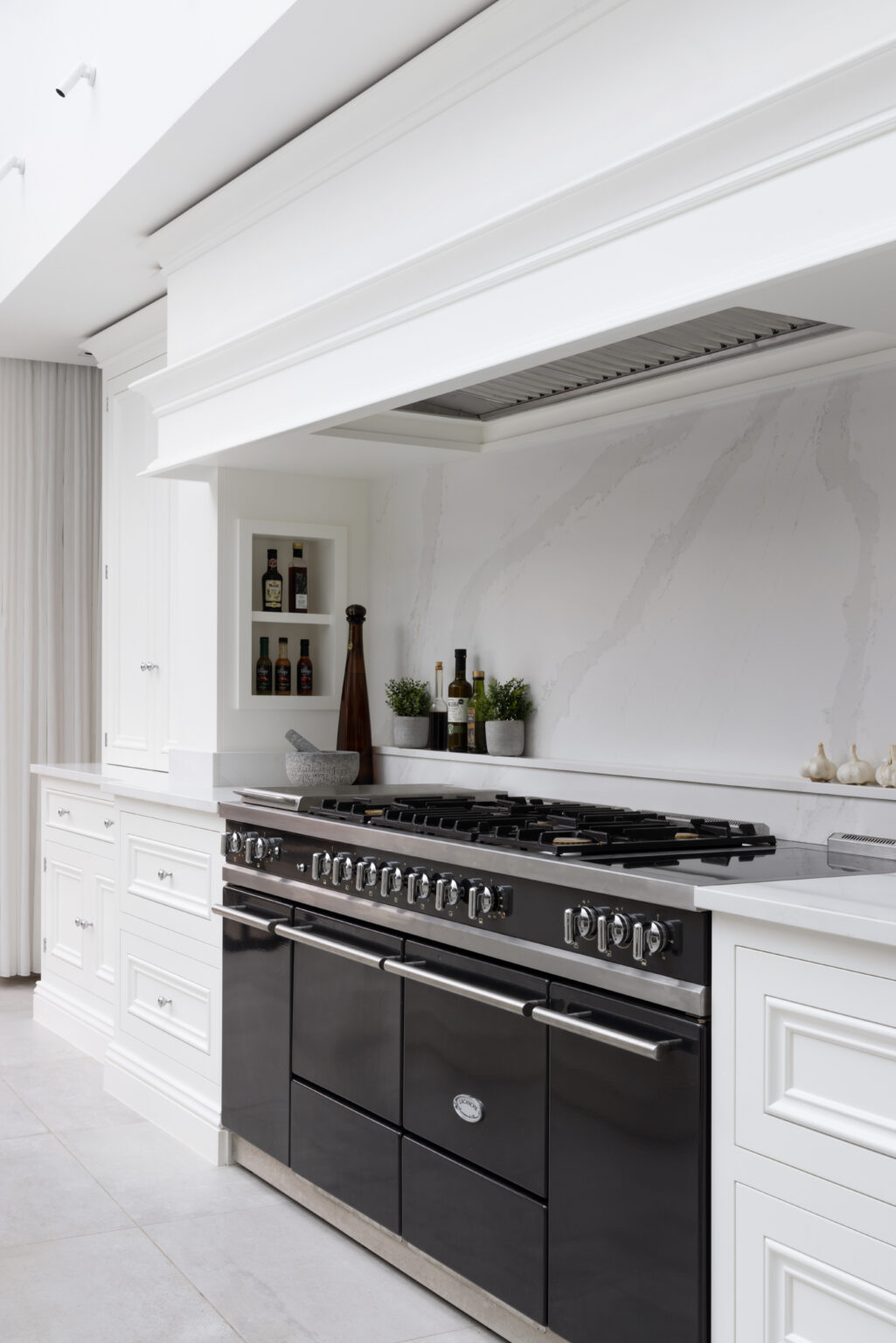 A black range cooker sits beneath a white panelled extractor hood, with bottles and herbs on the marbled splashback shelf