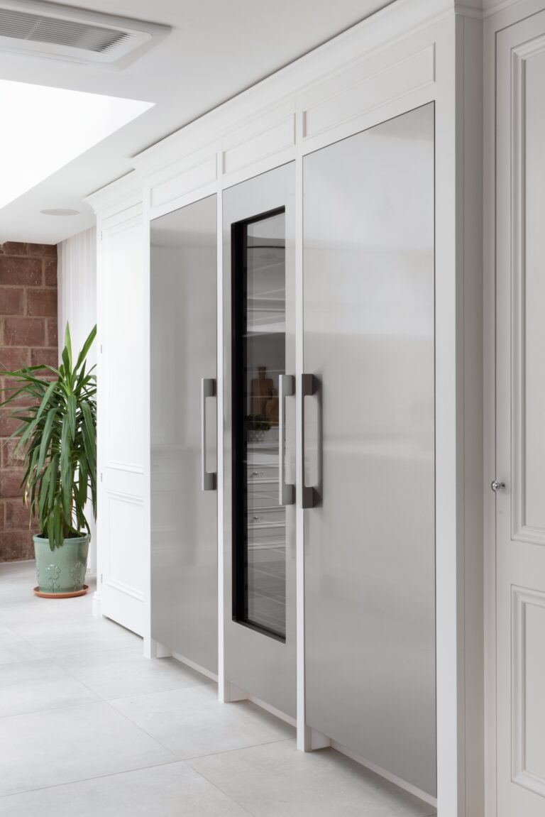 White kitchen cabinetry houses integrated fridge and wine cooler, with exposed sandstone wall visible through the doorway