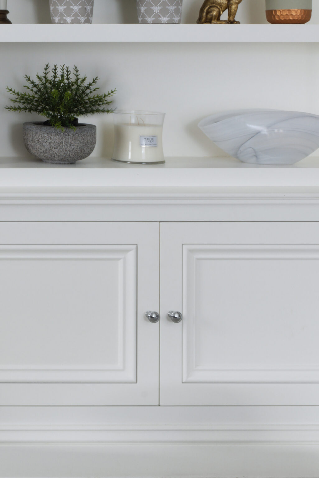 White panelled cabinet doors with chrome knobs, open shelving above displaying a plant, candle and bowl