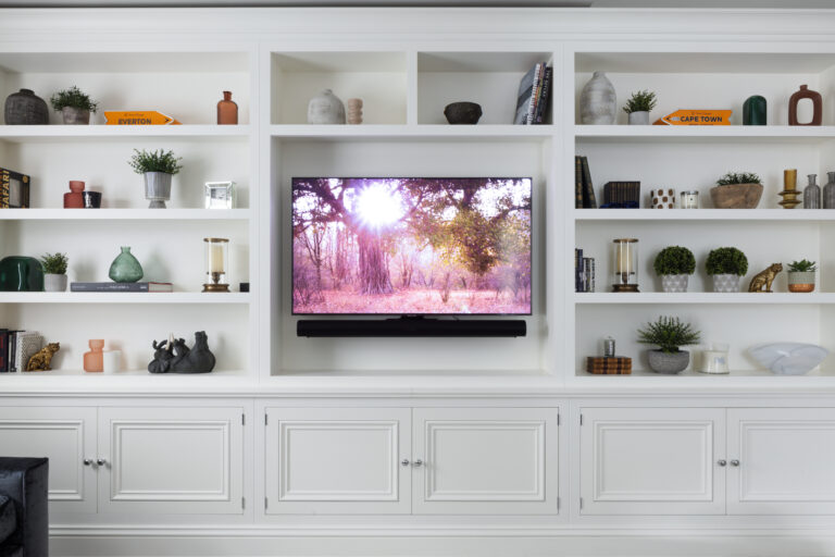 White fitted media lounge shelving with cupboards below and a wall-mounted television flanked by ornaments and books