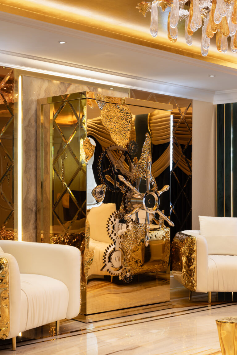 A gold-panelled lounge cabinet with sculptural mechanical details sits between cream armchairs beneath a crystal chandelier