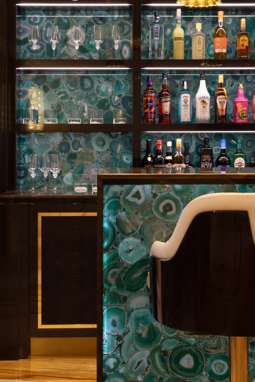 Backlit green agate bar with gold-edged shelving, glassware and spirit bottles above a cream leather stool