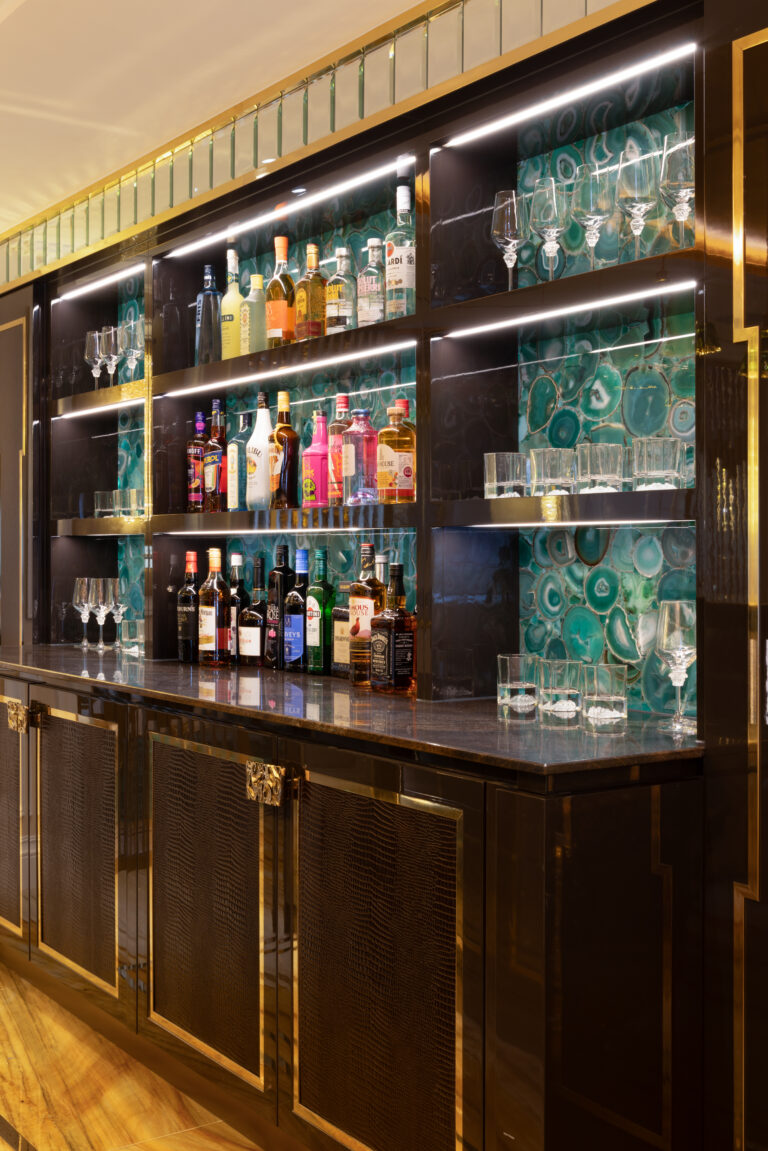 Bespoke bar with backlit green agate shelving, gold trim and dark glossy cabinetry displaying glassware and spirits