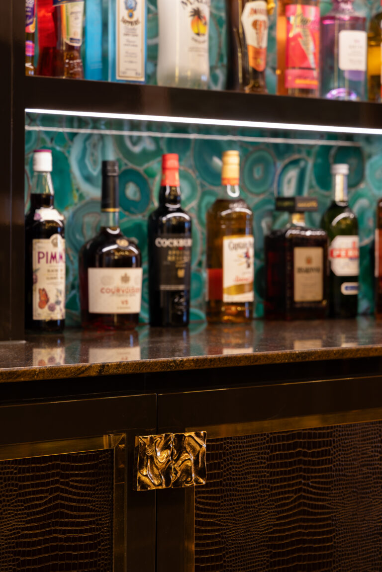 Bottles stand on a shelf against a green agate backsplash, above textured cabinetry with a sculpted gold handle