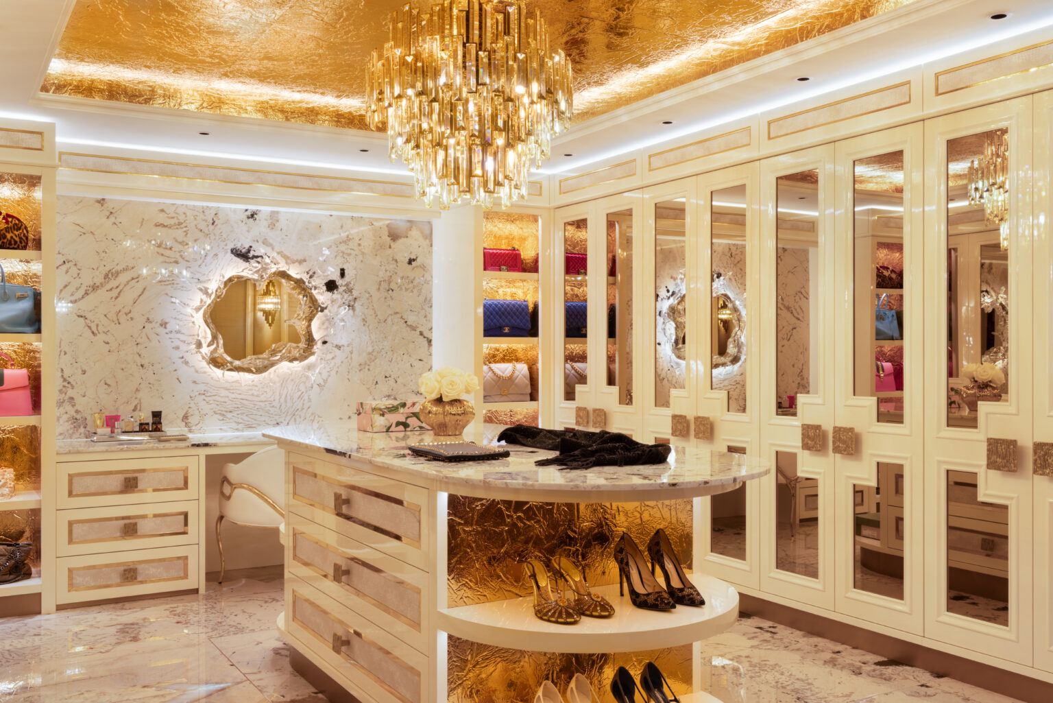 Dressing room with a gold ceiling, crystal chandelier, mirrored wardrobes and a central island displaying handbags and shoes