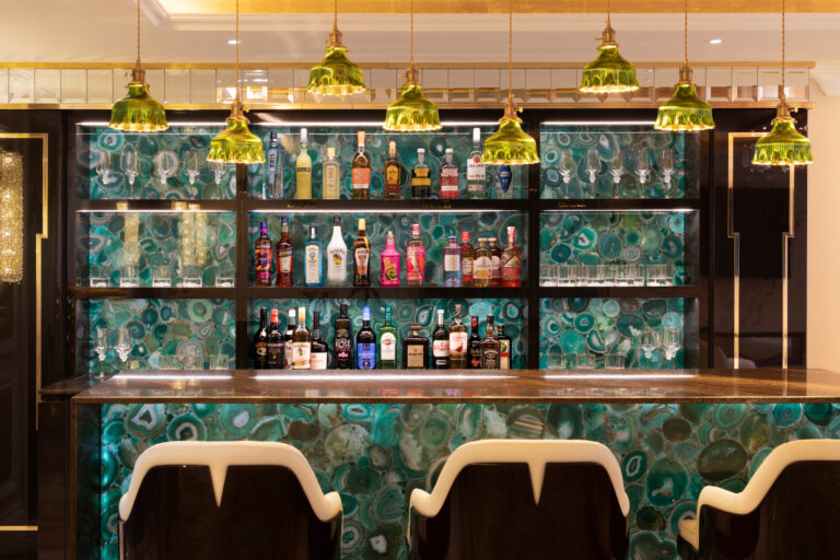 Home bar with green agate panelled shelving and front, gold pendant lights, bar stools and backlit glass shelves of bottles