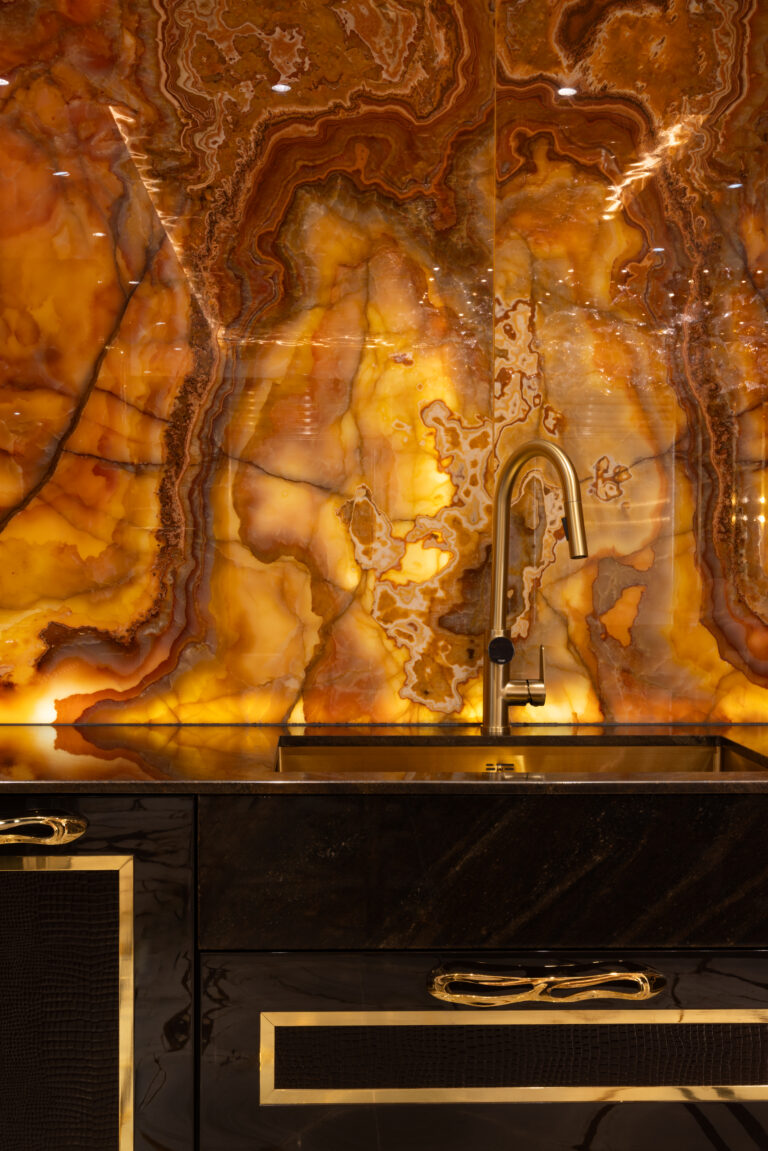 Backlit onyx splashback glows amber behind a gold sink and tap, above dark cabinetry with gold trim handles