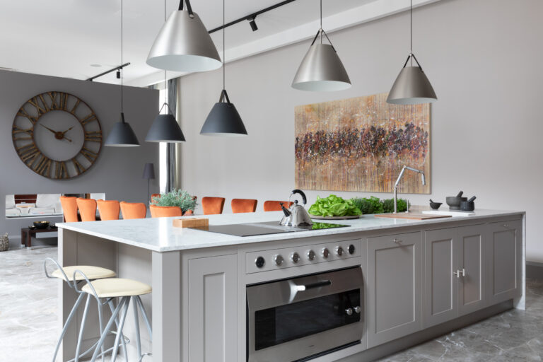 Large kitchen island with integrated oven and hob, stone worktop, pendant lighting and orange dining chairs beyond