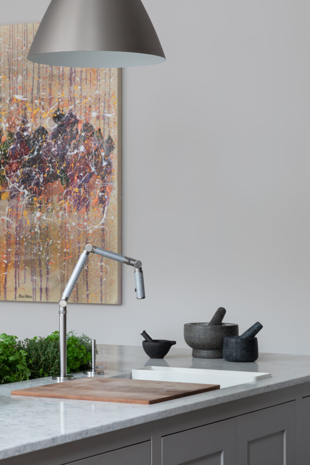 Kitchen worktop with articulated tap, pestle and mortars, herbs and chopping board beneath a pendant light and artwork