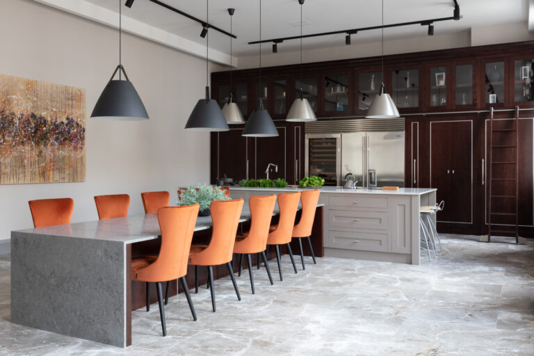 Long kitchen island with orange velvet chairs beneath dark cherry wood cabinetry and tall ceilings