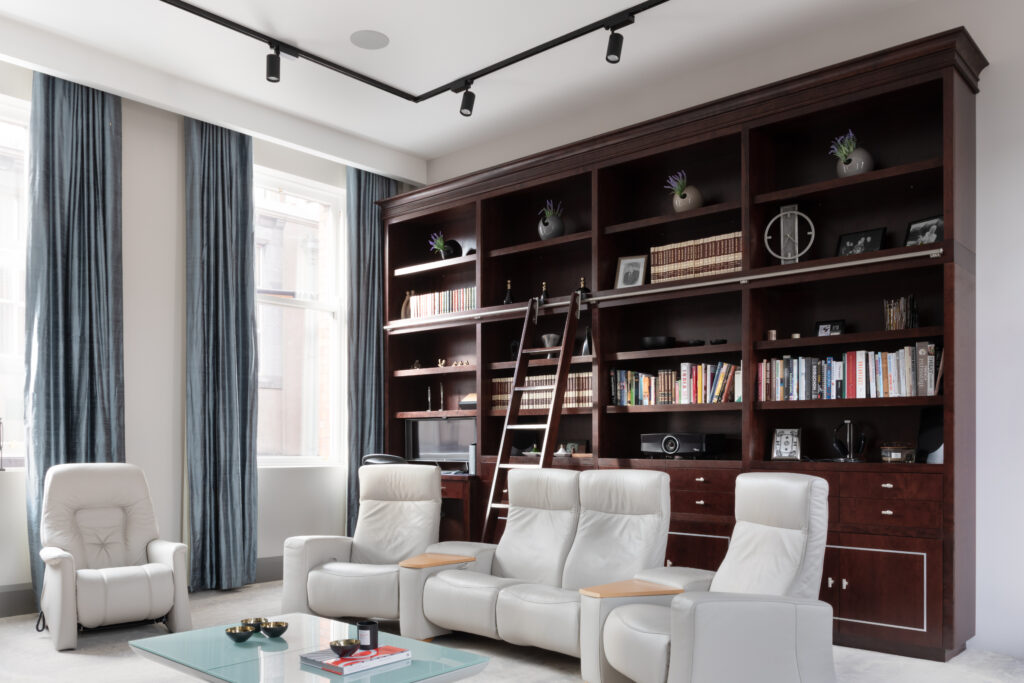 Floor-to-ceiling deep-stained cherry wood library wall with sliding ladder, facing pale leather recliners and a glass coffee table