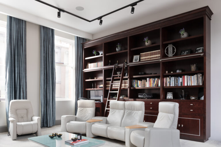 Floor-to-ceiling deep-stained cherry wood library wall with sliding ladder, facing pale leather recliners and a glass coffee table