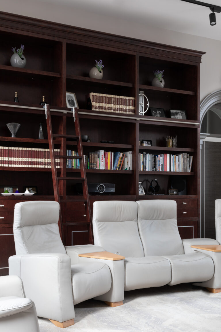 Deep-stained cherry wood library wall with sliding ladder, books and ornaments, above pale leather reclining cinema seats