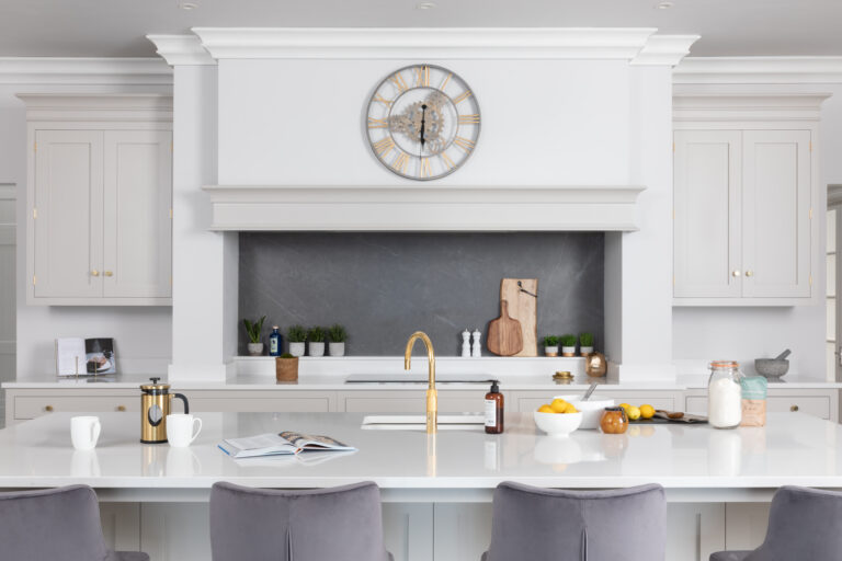Symmetrical Cornforth White cabinetry frames the sink area, with a gold tap and grey velvet stools at the island